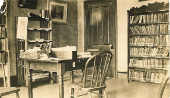 Black and white photo of the library. There are a couple book cases and a desk with a chair.
