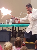 Photograph of a man in a lap coat blowing into a tube and it appears fire is coming out the other end.